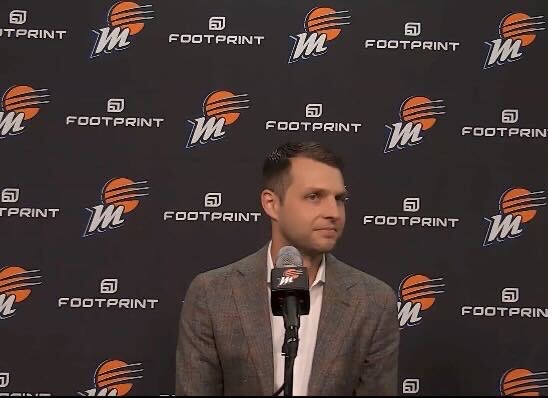 Incoming Phoenix Mercury general manager Nick U'Ren sits at the podium, looking to his left, as he takes questions from reporters. A backdrop featuring many Mercury logos is visible behind him.