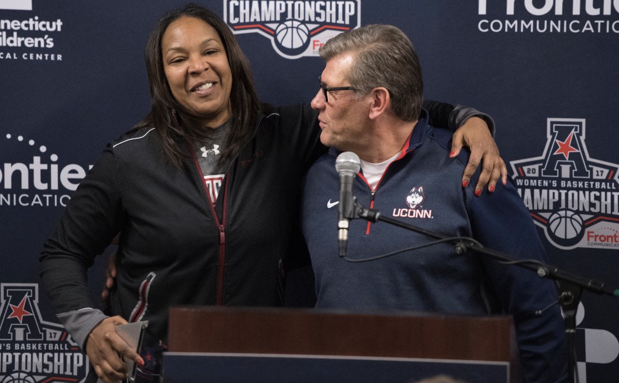 In a photo from 2017, then-Temple head coach Tonya Cardoza puts her arm around UConn head coach Geno Auriemma against a backdrop of American Athletic Conference Tournament branding.