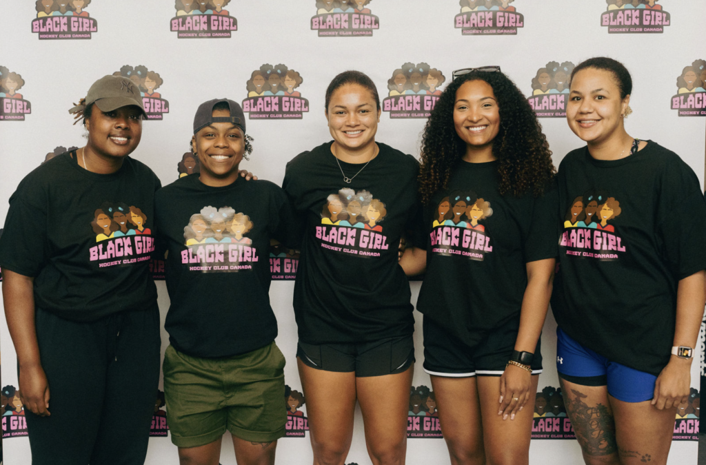 Five participants in the Black Girl Hockey Club's first-ever camp are wearing black t-shirts featuring the club's logo against a white backdrop featuring the same logo.