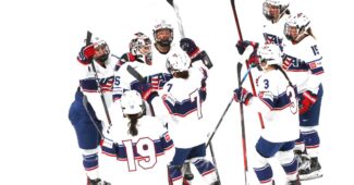 Members of the U18 USA Hockey team celebrate by lifting their hockey sticks in the air in a huddle. They are wearing the traditional red, white and blue USA Hockey uniforms.