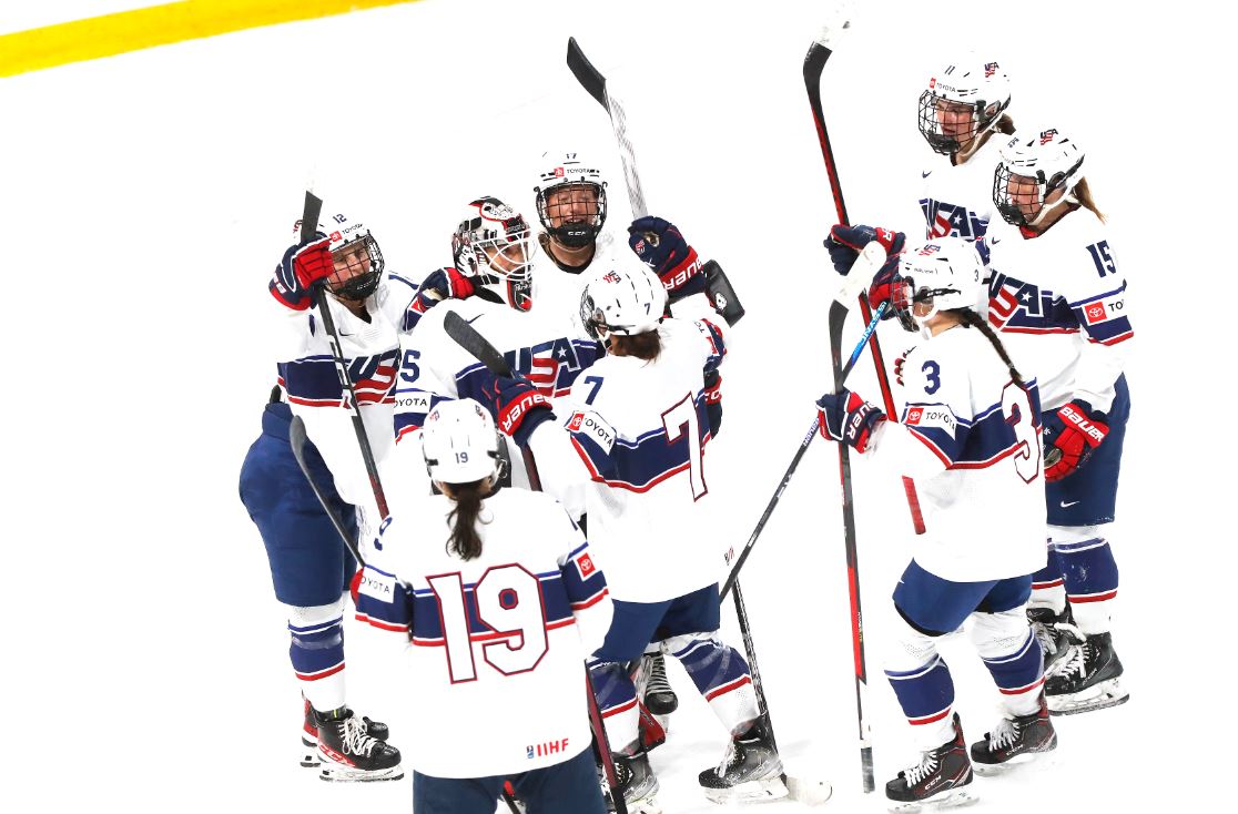 Members of the U18 USA Hockey team celebrate by lifting their hockey sticks in the air in a huddle. They are wearing the traditional red, white and blue USA Hockey uniforms.
