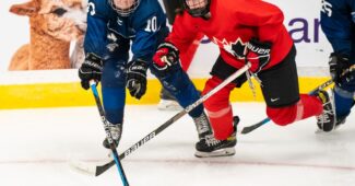Sienna D’Alessandro wearing a red Team Canada jersey fights on the ice for position to get the puck against an opposing player in a blue jersey during Team Canada's U18 Selection Camp.