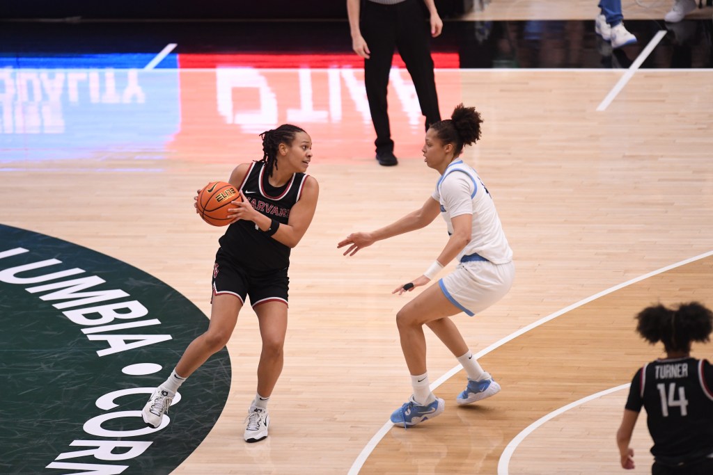Harvard's McKenzie Forbes holds the ball near the Ivy League Tournament logo at center court as Columbia's Kaitlyn Davis defends.
