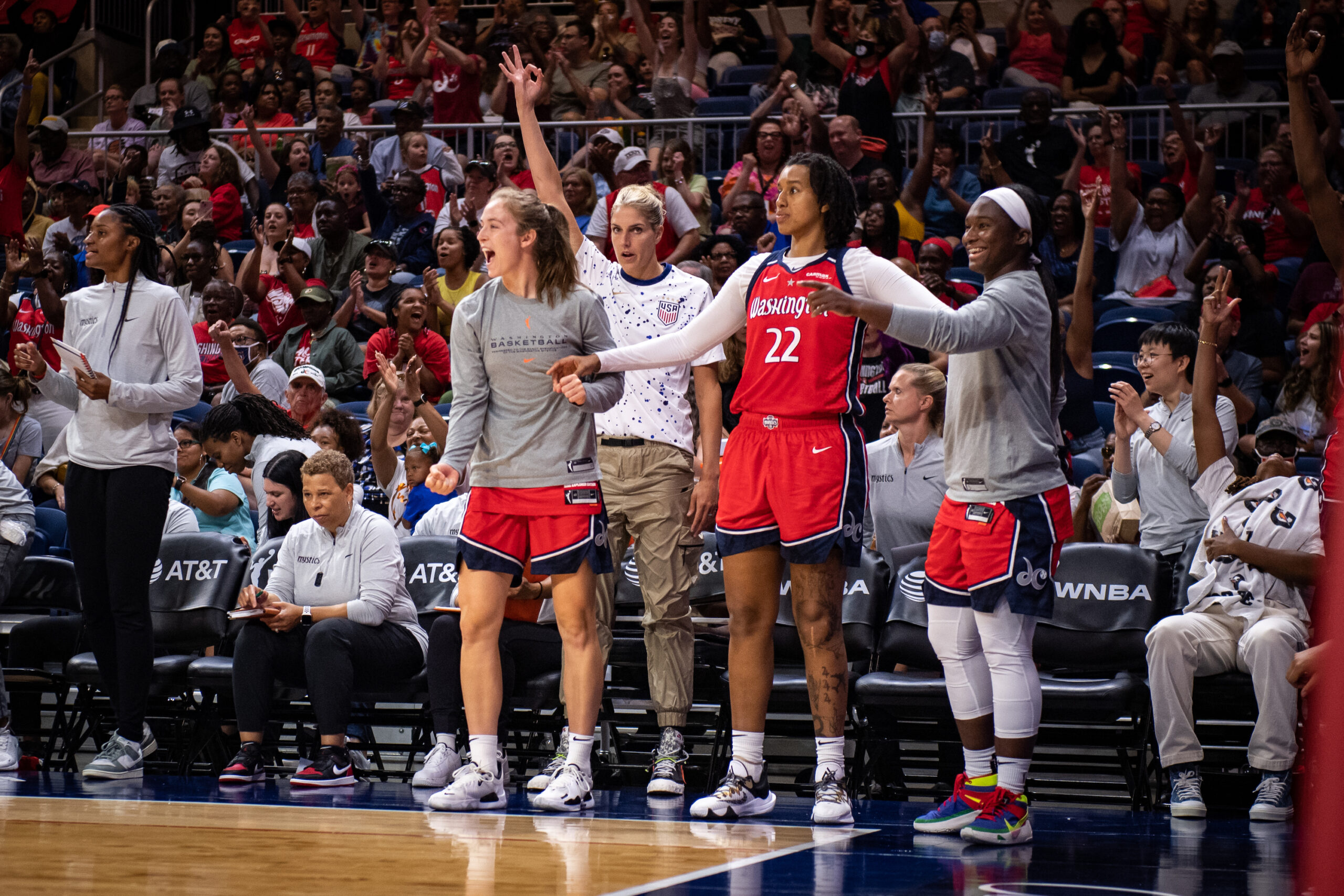 Washington Mystics players Abby Meyers, Cyesha Goree, Linnae Harper and Elena Delle Donne stand and celebrate on the bench during a game. Meyers is shouting, and Delle Donne has her right hand in the air making the 3-point sign.