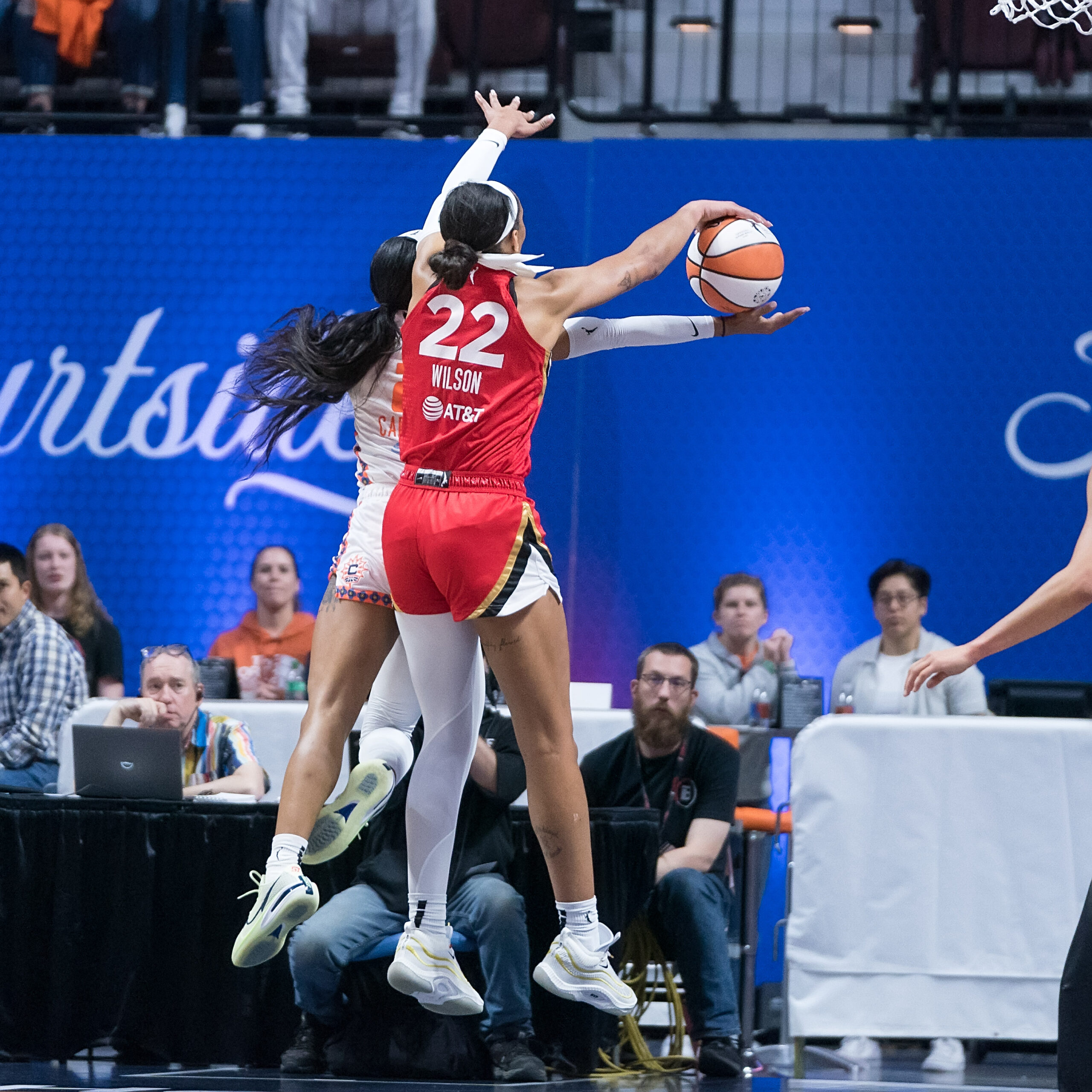 As Connecticut Sun guard DiJonai Carrington releases a layup with her arm parallel to the floor, Las Vegas Aces forward A'ja Wilson blocks it. Both players are shown in mid-air with their backs to the camera, their hands sandwiching the ball.