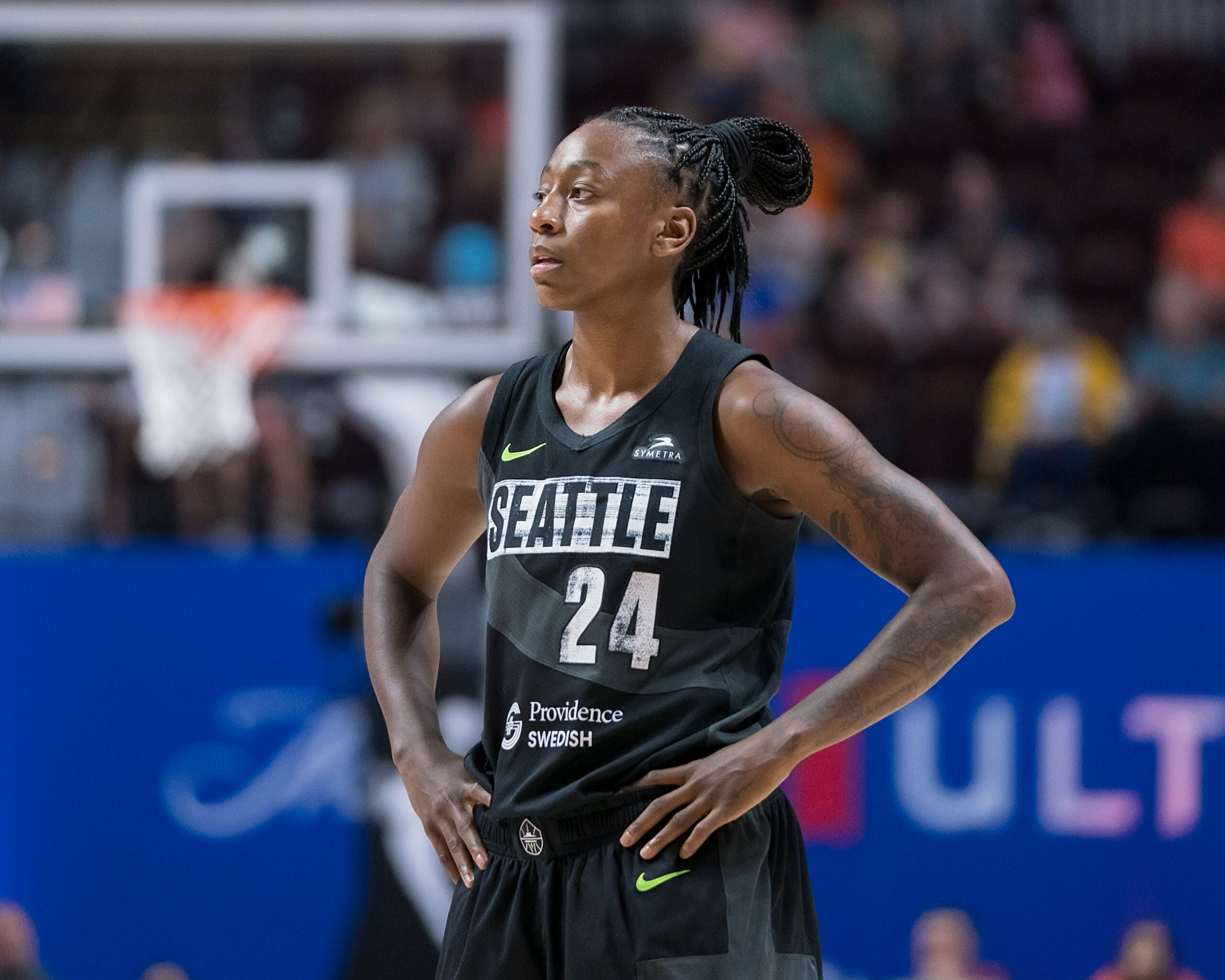 Jewell Loyd stands with a basketball hoop in the distance, wearing Seattle's black uniform, with her hands on hips.
