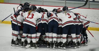 The RMU women's hockey team huddles up. The team is on the ice in uniform, each player holding a hockey stick.