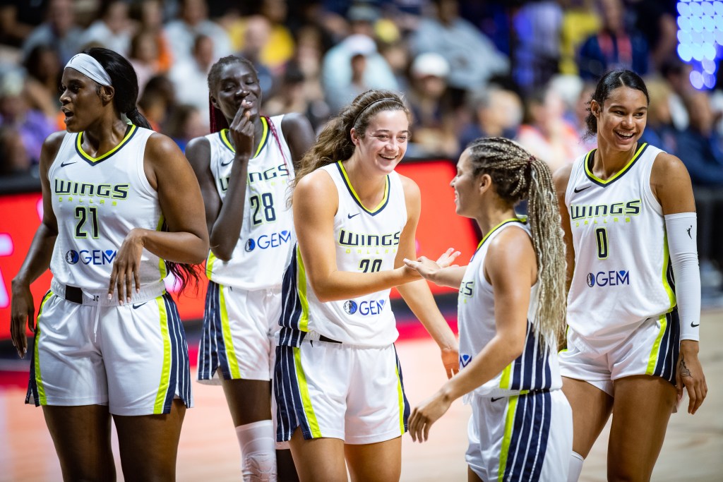 Five Dallas Wings players cluster together on the court. Awak Kuier and Satou Sabally smile in the background, while Maddy Siegrist and Veronica Burton high-five.