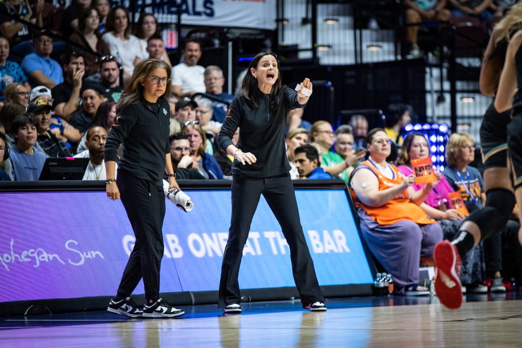 New York Liberty head coach Sandy Brondello holds her hand out and shouts instructions from the sideline during a game. Head athletic trainer Terri Acosta stands next to her and watches.