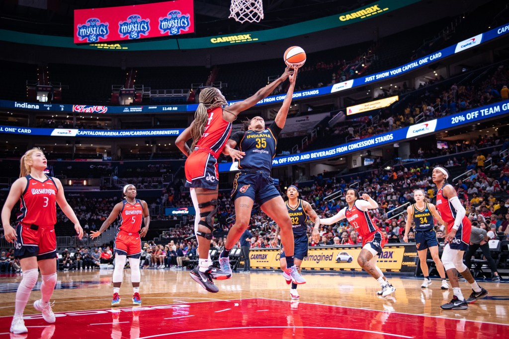 Washington Mystics center Queen Egbo reaches her right arm across the body of Indiana Fever guard Victoria Vivians to block Vivians' left-handed layup attempt.