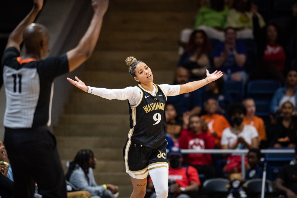 Washington Mystics guard Natasha Cloud extends her arms to the side and tilts her head slightly to the right, celebrating a made 3-pointer. The nearest official holds two arms up to signal that the 3-pointer counts.