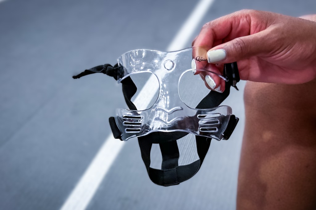 A close-up photo of a clear protective face mask. It has adjustable black straps and small holes in the mask to allow for ventilation.