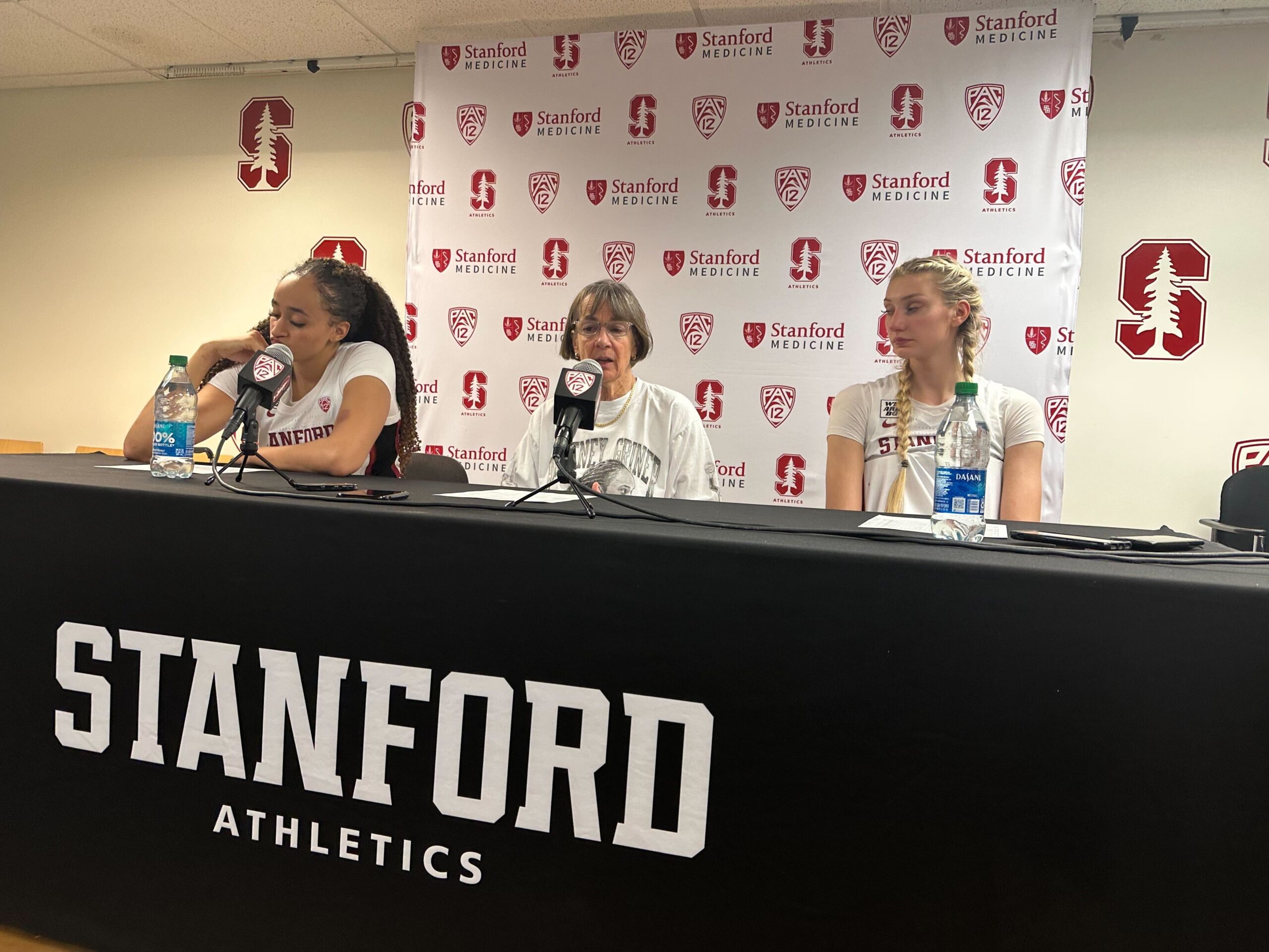 Stanford head coach Tara VanDerveer sits at the podium and speaks at a press conference. Two of her players are on either side of her, and a backdrop of Stanford and Pac-12 logos is displayed behind them.