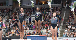 Shilese Jones, Simone Biles and Leanne Wong stand on the podium and hold bouquets of flowers aloft in their right hands.
