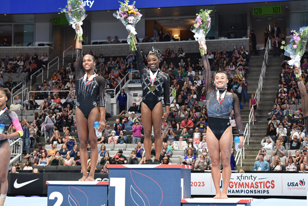 Shilese Jones, Simone Biles and Leanne Wong stand on the podium and hold bouquets of flowers aloft in their right hands.