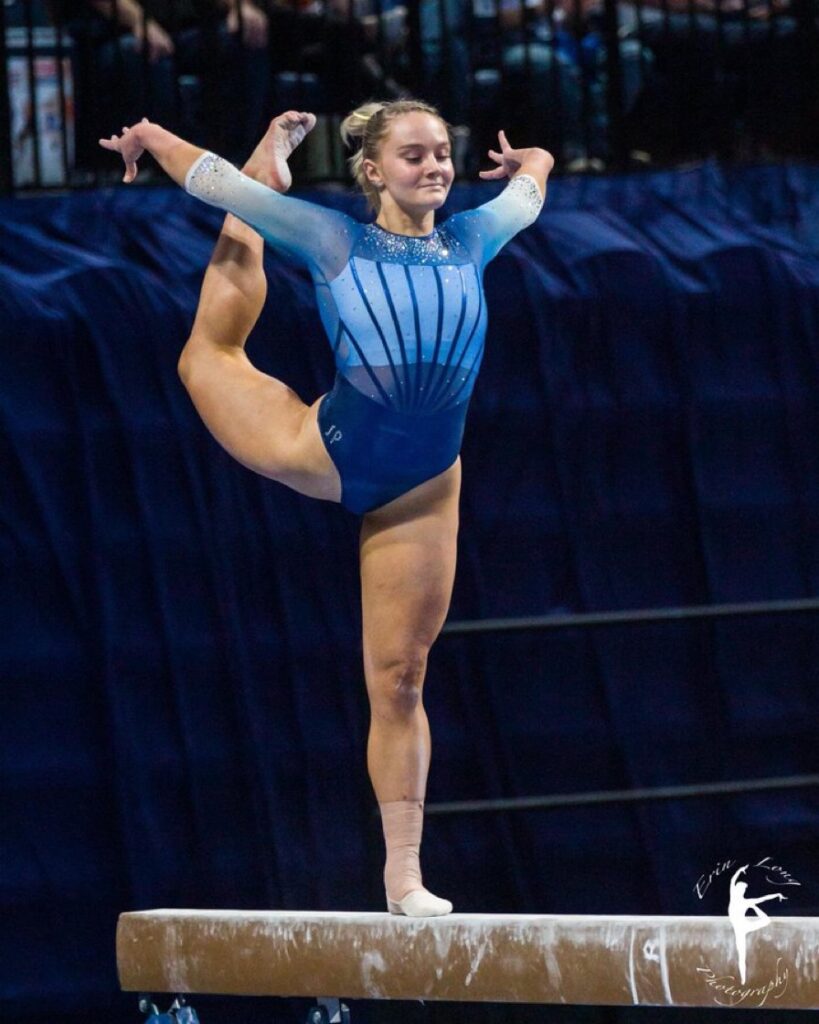 Riley McCusker stands on the balance beam on her left leg. Her right leg curves behind her and up, and her arms extend behind her.