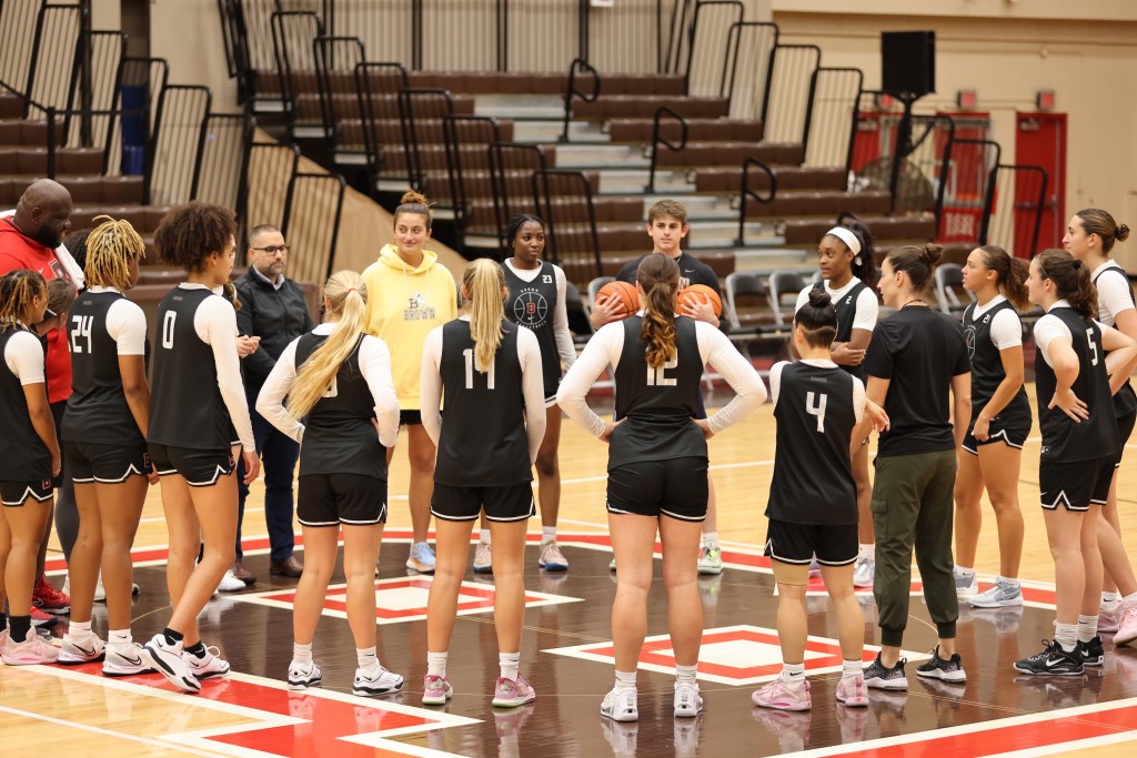 The Brown women's basketball team stands in a circle on the large "B" logo in the middle of its home court.