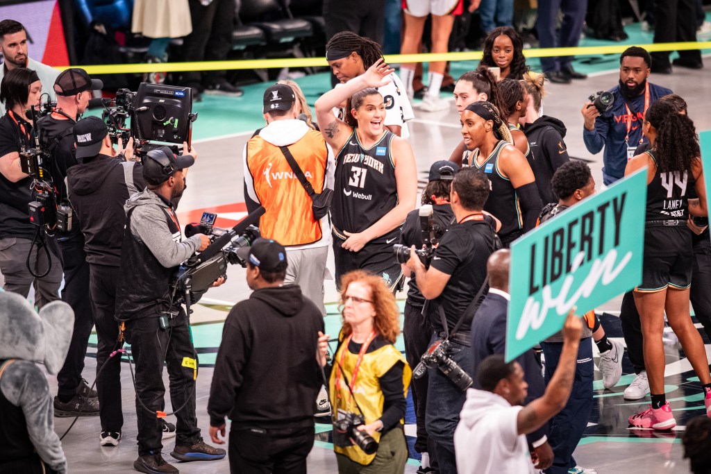 A crowd of photographers surrounds New York Liberty players on the court after a game. Forward Stefanie Dolson is in the middle and waves with her right hand to someone off camera.