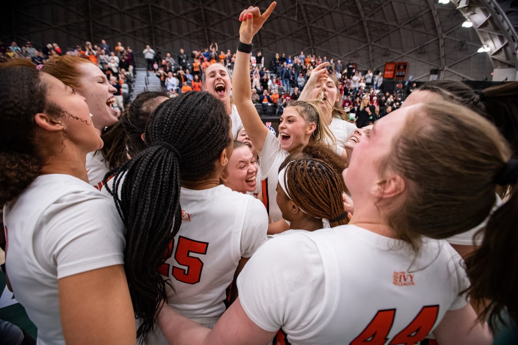 Princeton players hug each other and scream in celebration after winning the Ivy League Tournament title. Forward Ellie Mitchell is shown in the center of the photo, beaming and holding her right pointer finger in the air.
