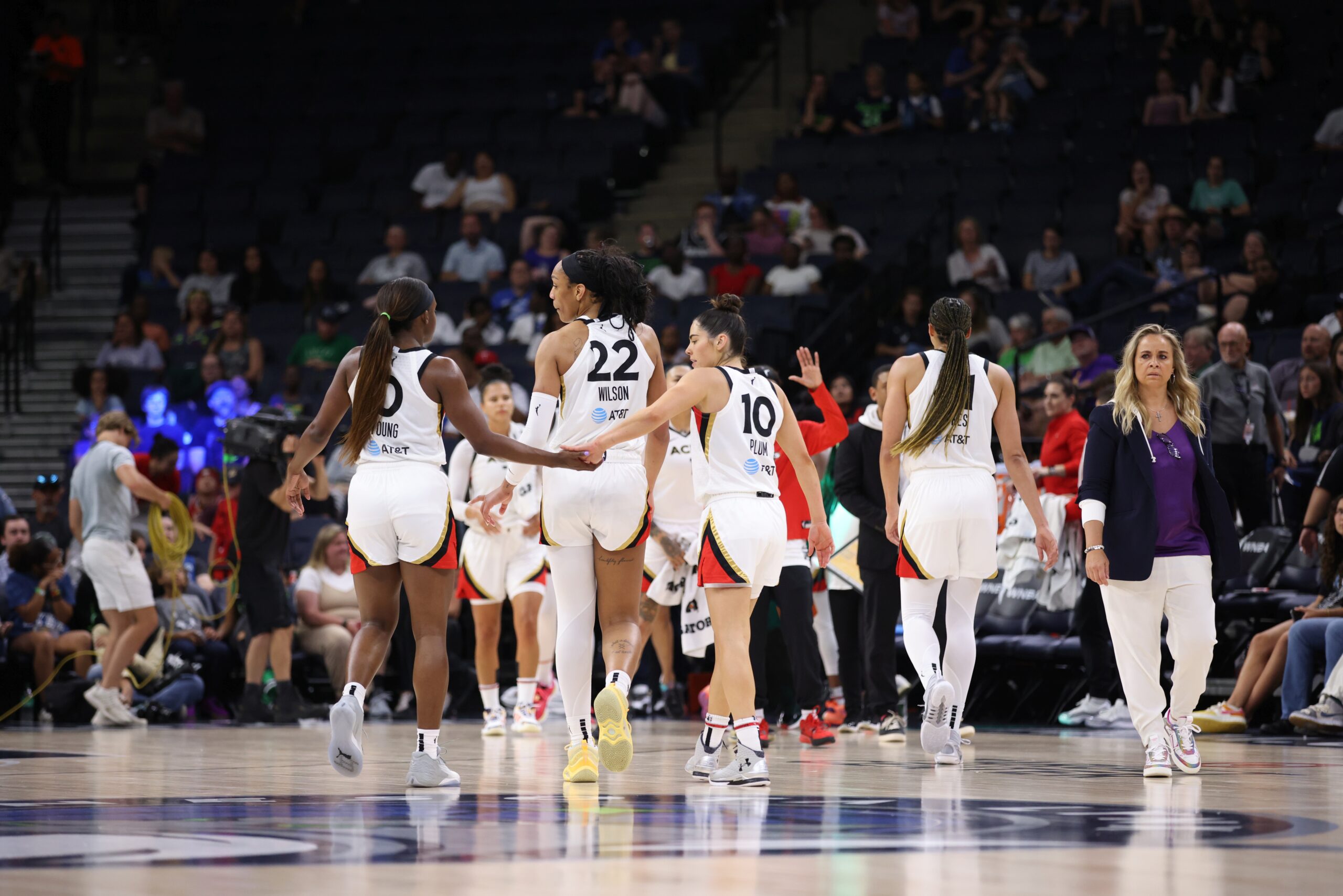 Las Vegas Aces players including Jackie Young, A'ja Wilson and Kelsey Plum walk merrily down the court toward the end of their team's bench. Head coach Becky Hammon stands on the side, gazing toward the middle of the court in mild bewilderment.
