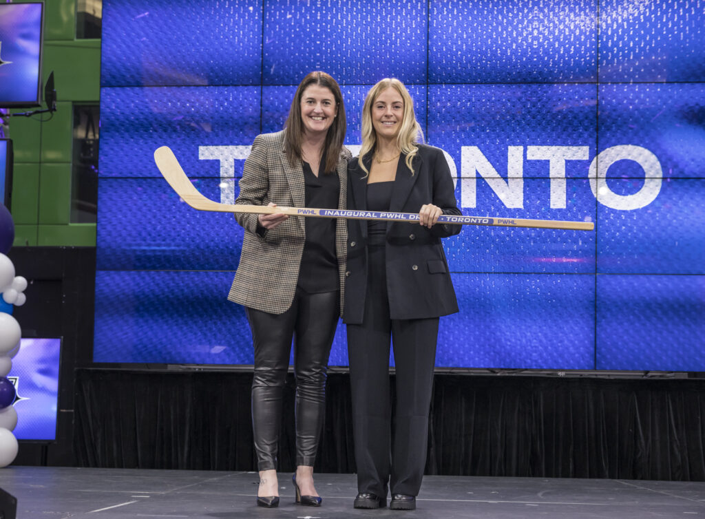 PWHL draftee Rebecca Leslie stands holding a hockey stick alongside PWHL leader wearing black blazer and black dress pants.