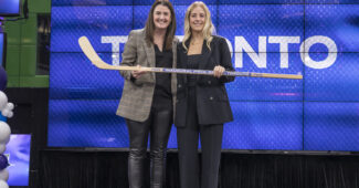 PWHL draftee Rebecca Leslie stands holding a hockey stick alongside PWHL leader wearing black blazer and black dress pants.