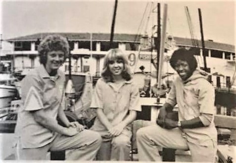 In a black and white photo, San Francisco Pioneers teammates Cindy Haugejorde, Molly (Bolin) Kazmer and Cardte Hicks pose for a photograph in the team warm-up uniforms. They are posing against a backgroup of a boat and are seemingly kneeling on a boat dock.