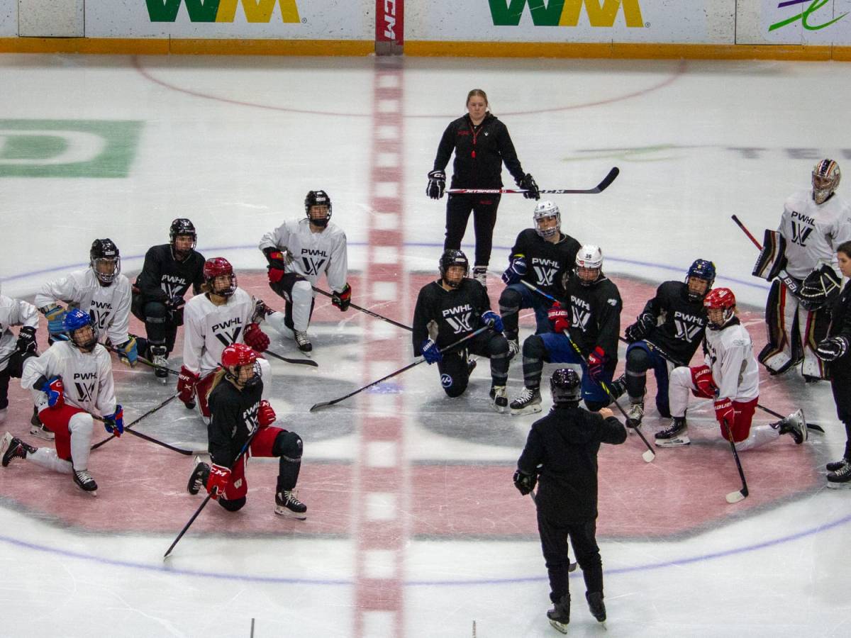 Hockey players in PWHL jerseys take a knee at centre ice infront of coach Carla MacLeod.