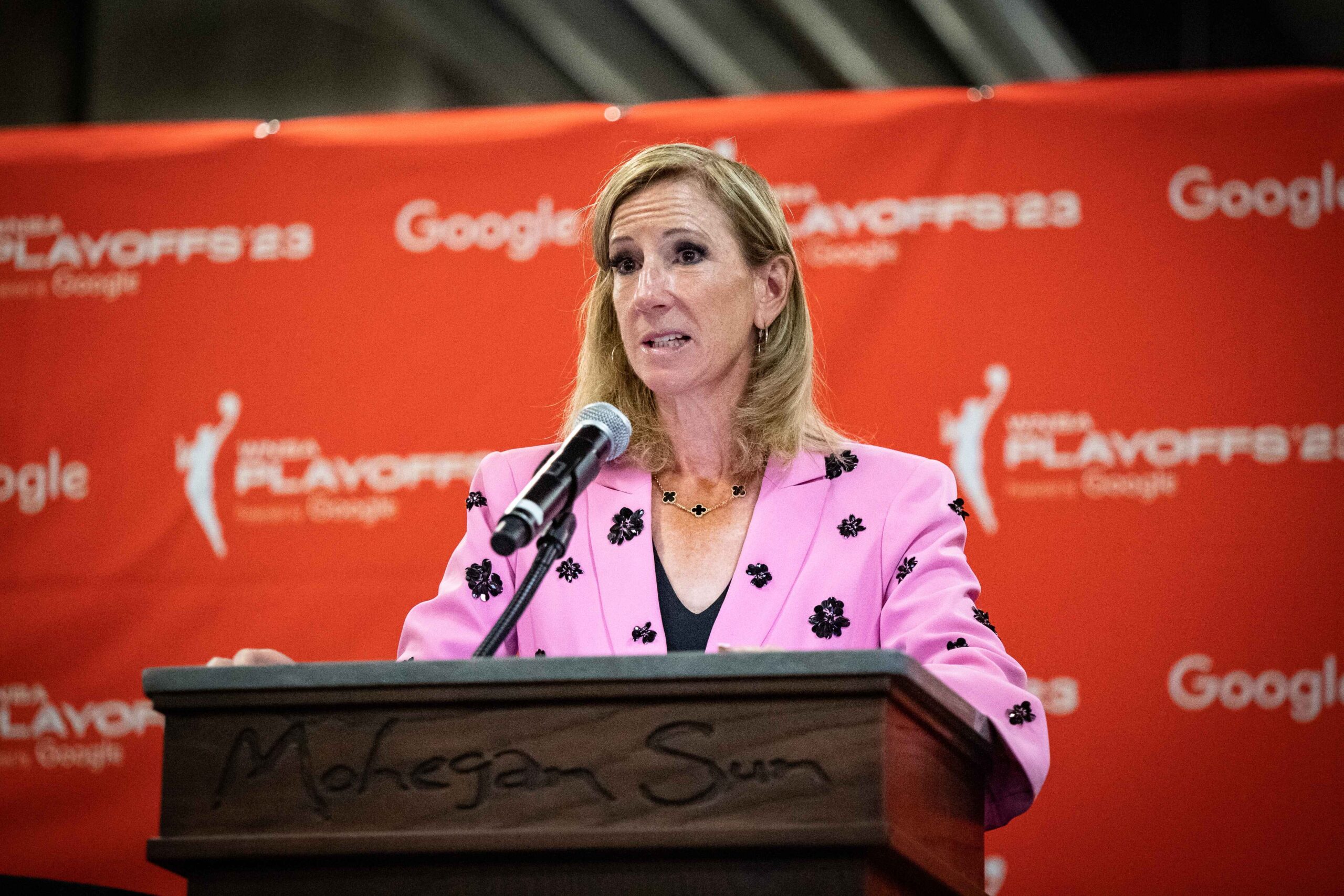 WNBA Commissioner Cathy Engelbert stands at a wooden podium and speaks with the media ahead of a 2023 playoff game. An orange backdrop featuring the WNBA playoffs and Google logos is behind her.