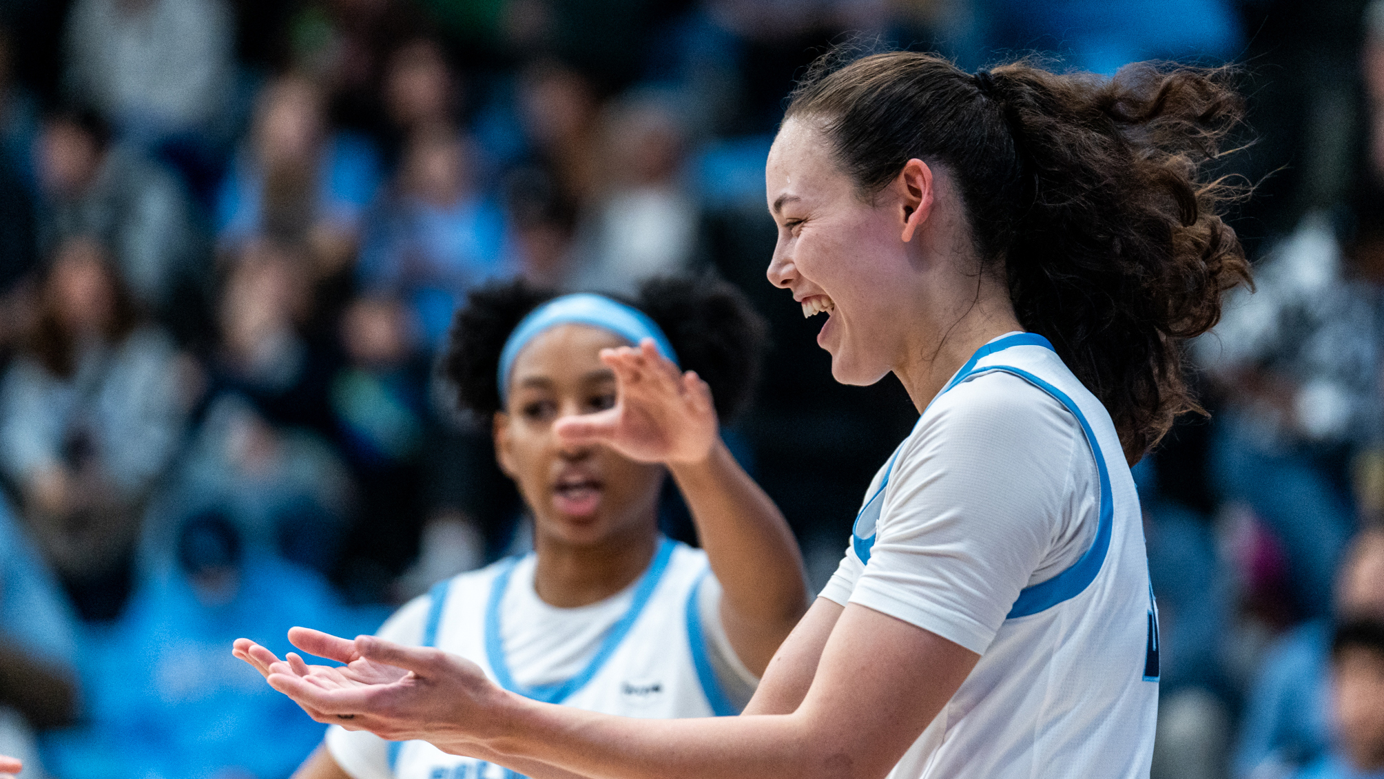 Columbia guard Abbey Hsu is shown in profile, smiling with her hands extended in front of her and her palms up. Guard Perri Page is walking up to her and extending her left arm to complete the high five.