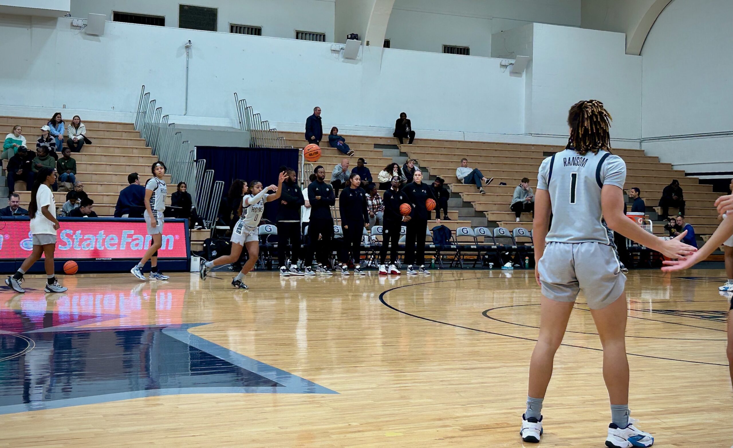 Georgetown graduate assistant Bella Alarie holds a ball under her left arm and stands with several other members of the Hoyas' staff on the sidelines, watching players warm up.