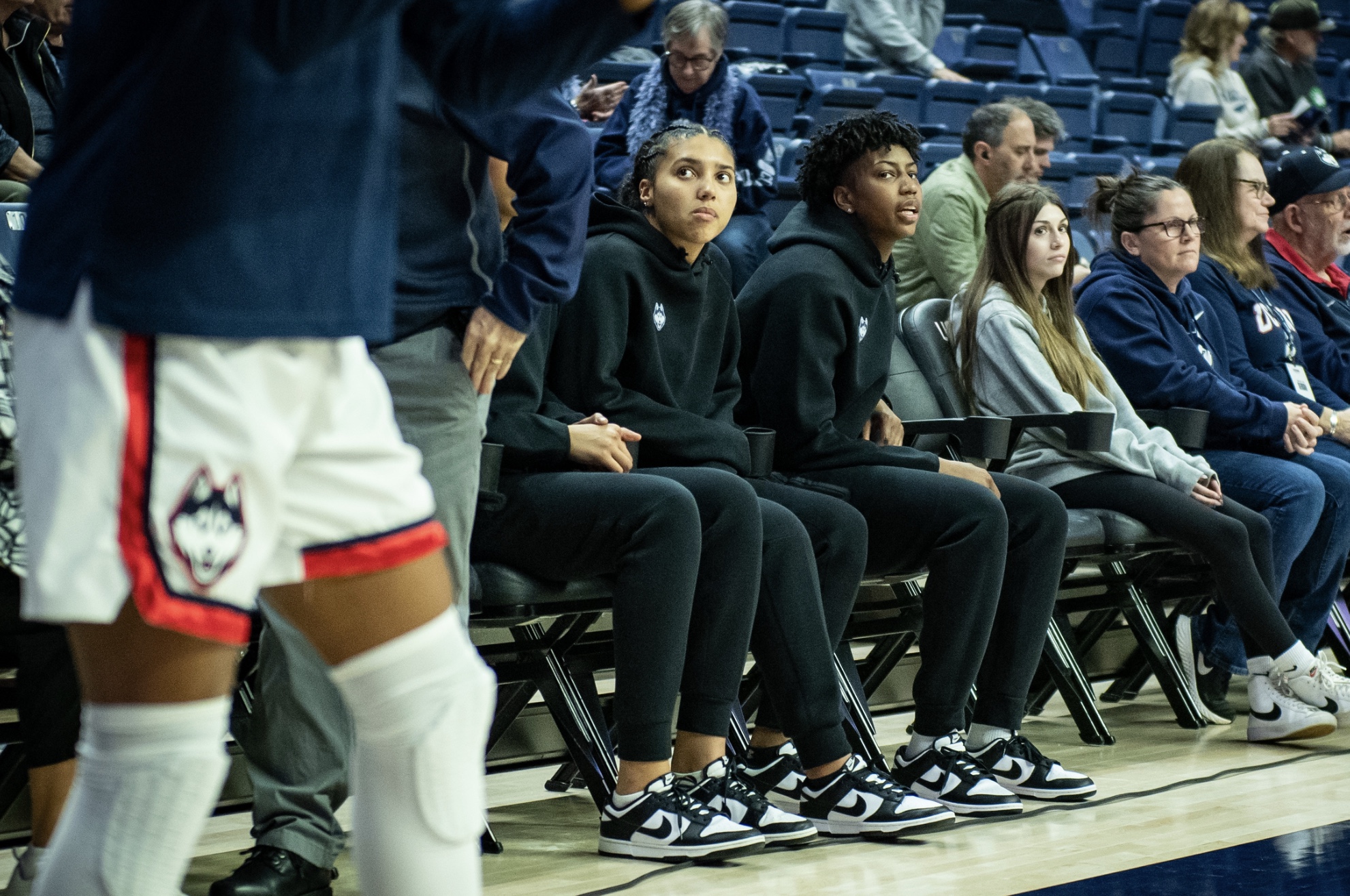 UConn junior Azzi Fudd watches from the bench while teammates warm up. She is wearing UConn's black warm up uniform.