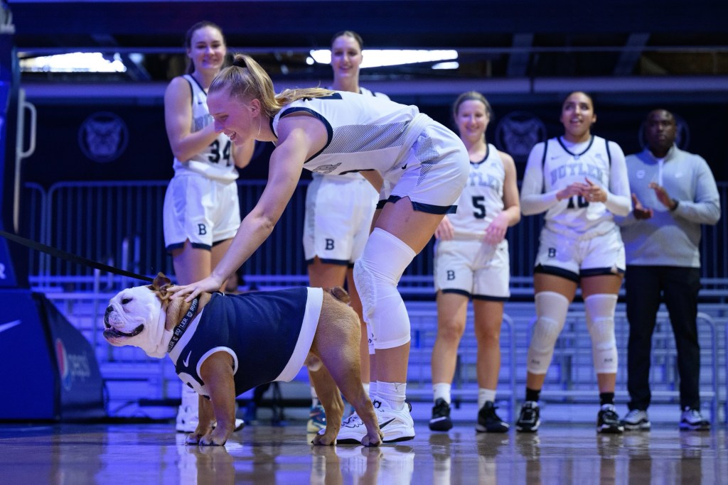 Butler sophomore Jordan Meulemans bends over to pet mascot Blue, a bulldog, who is wearing a blue sweater, as her teammates watch.