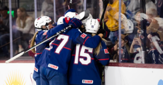 Members of the Team USA women's hockey team gather to celebrate while on the ice and wearing navy blue jerseys.