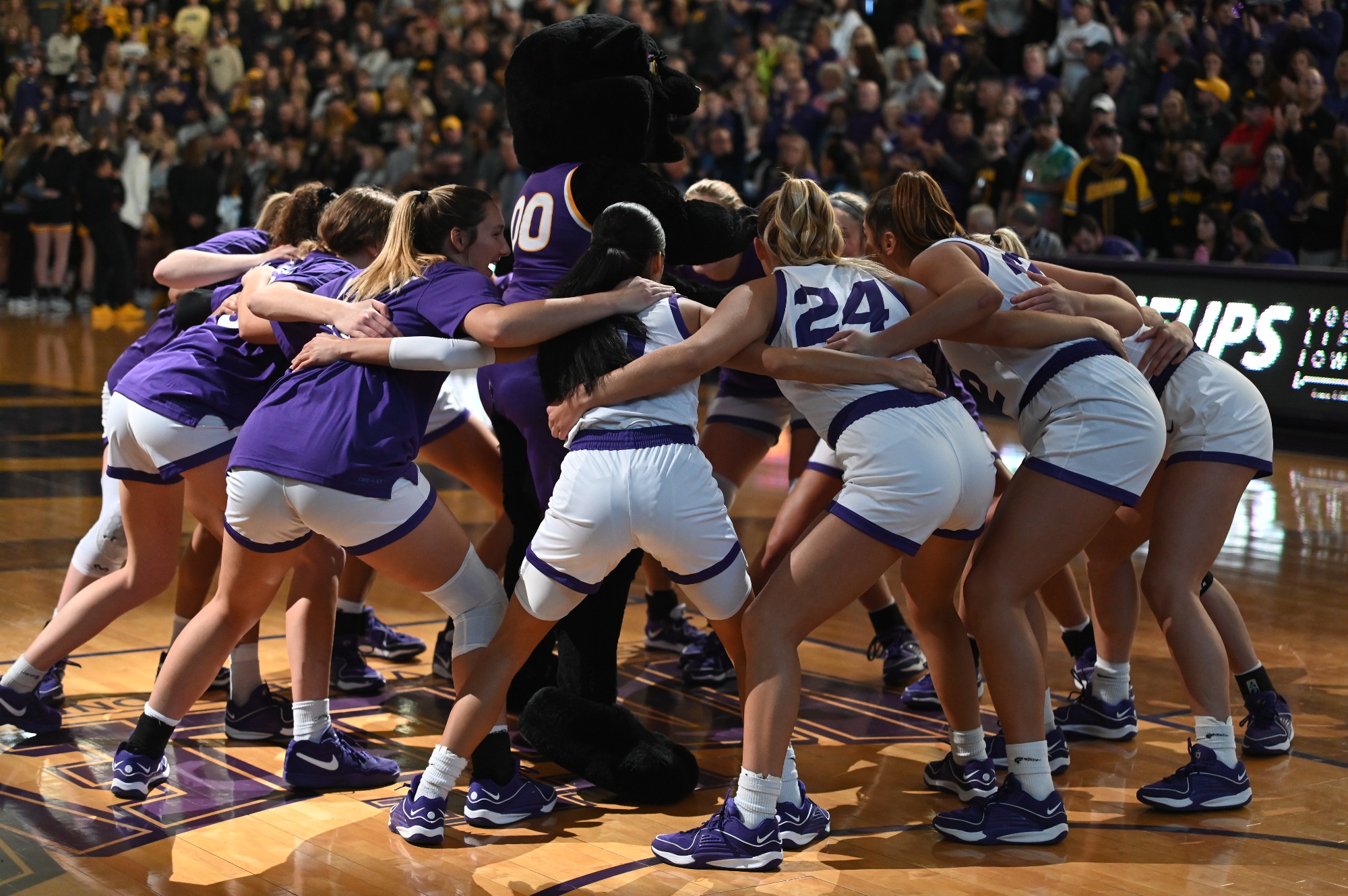UNI Panthers players huddle in a circle at center court, the lights dimmed, with their arms around each other as they prepare for a home game against the Iowa Hawkeyes