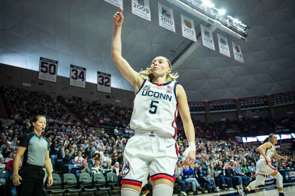 UConn guard Paige Bueckers is pictured doing the "and-one" sign following a drive to the basket. She is wearing UConn's home white jerseys against the backdrop of UConn's championship banners.