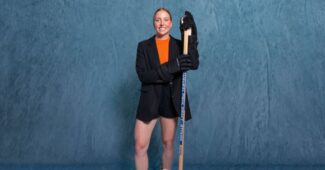 Olympian Ashton Bell is pictured with a hockey stick in front of a blue photo backdrop.