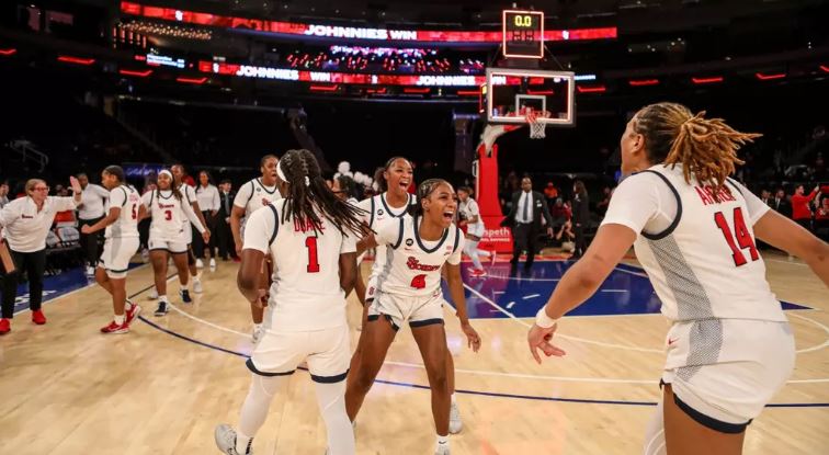 St. John's players celebrate together on the court at MSG following a win over Villanova. They are wearing white and red home uniforms.