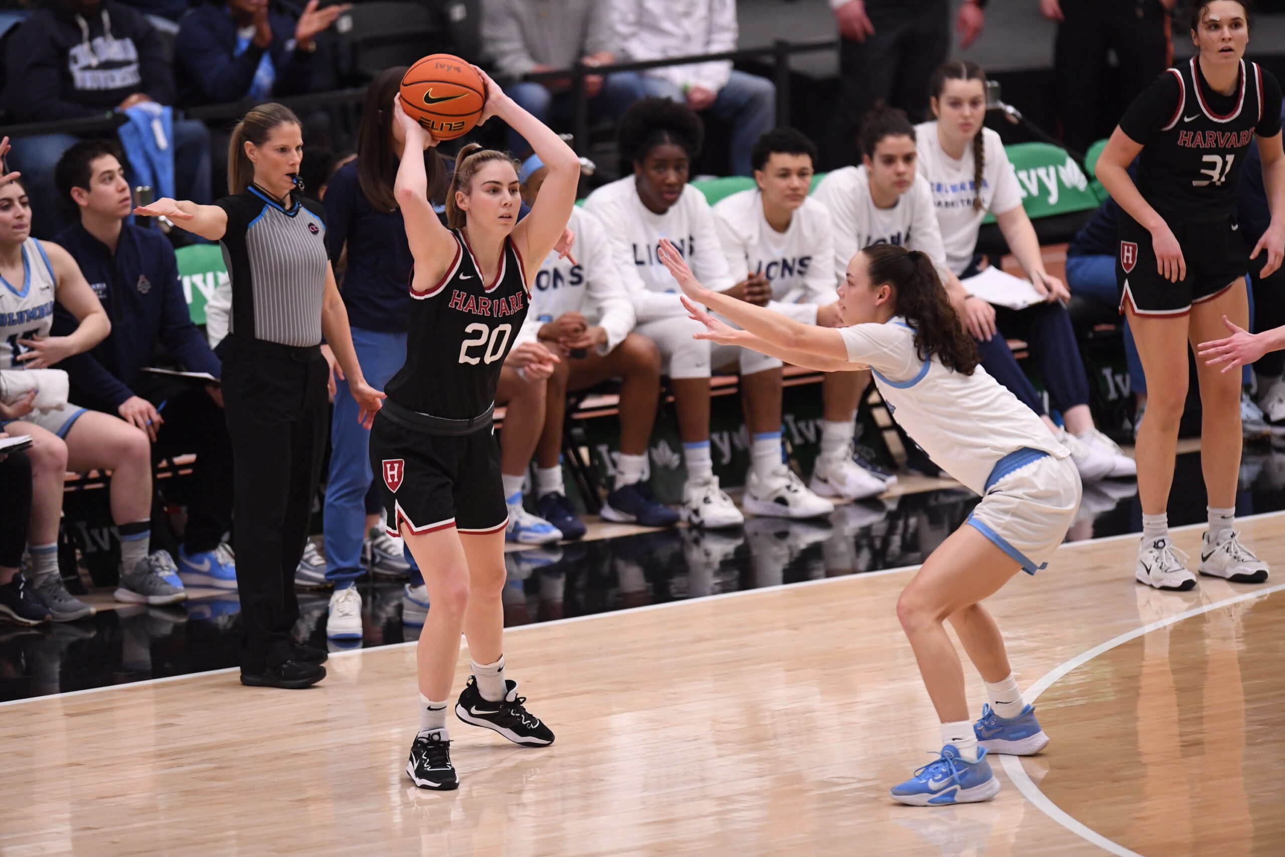 Harvard guard Lola Mullaney stands on the perimeter near the Columbia bench and pulls the ball behind her head with both hands to pass toward the center of the court. Columbia guard Abbey Hsu stands on the 3-point line with her hands out to pressure Mullaney.