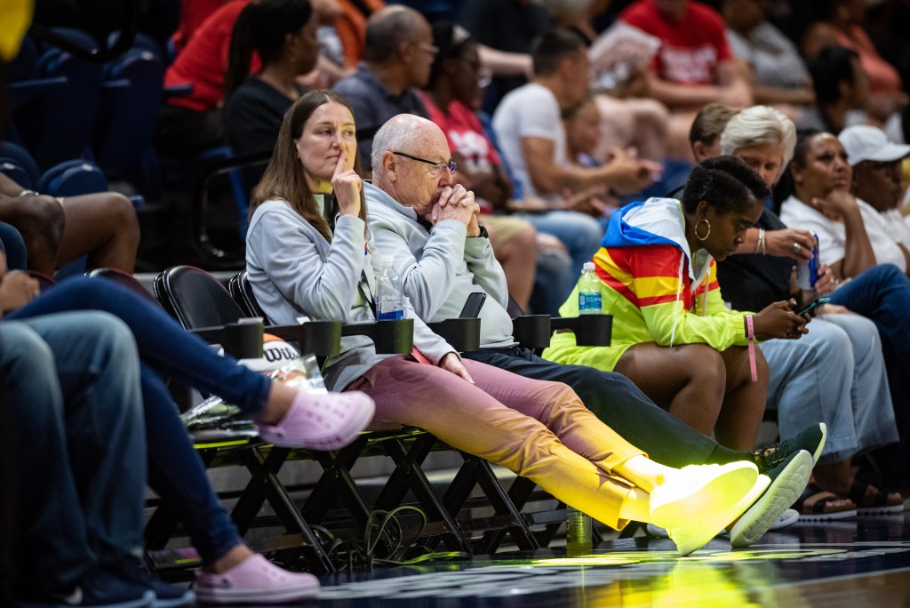 Sitting courtside during a game, Washington Mystics general manager Mike Thibault clasps his fingers and presses them to his chin. Assistant general manager Maria Giovannetti sits next to him with her right index finger pressed against her lips.