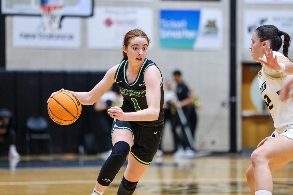 Dartmouth guard Nina Minicozzi looks up as she dribbles the ball with her right hand. She has her left arm bent and in front of her to protect the ball for a Bryant defender.