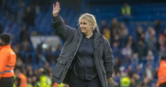 USWNT head coach Emma Hayes is pictured wearing a large overcoat and waving her right hand to a stadium crowd.
