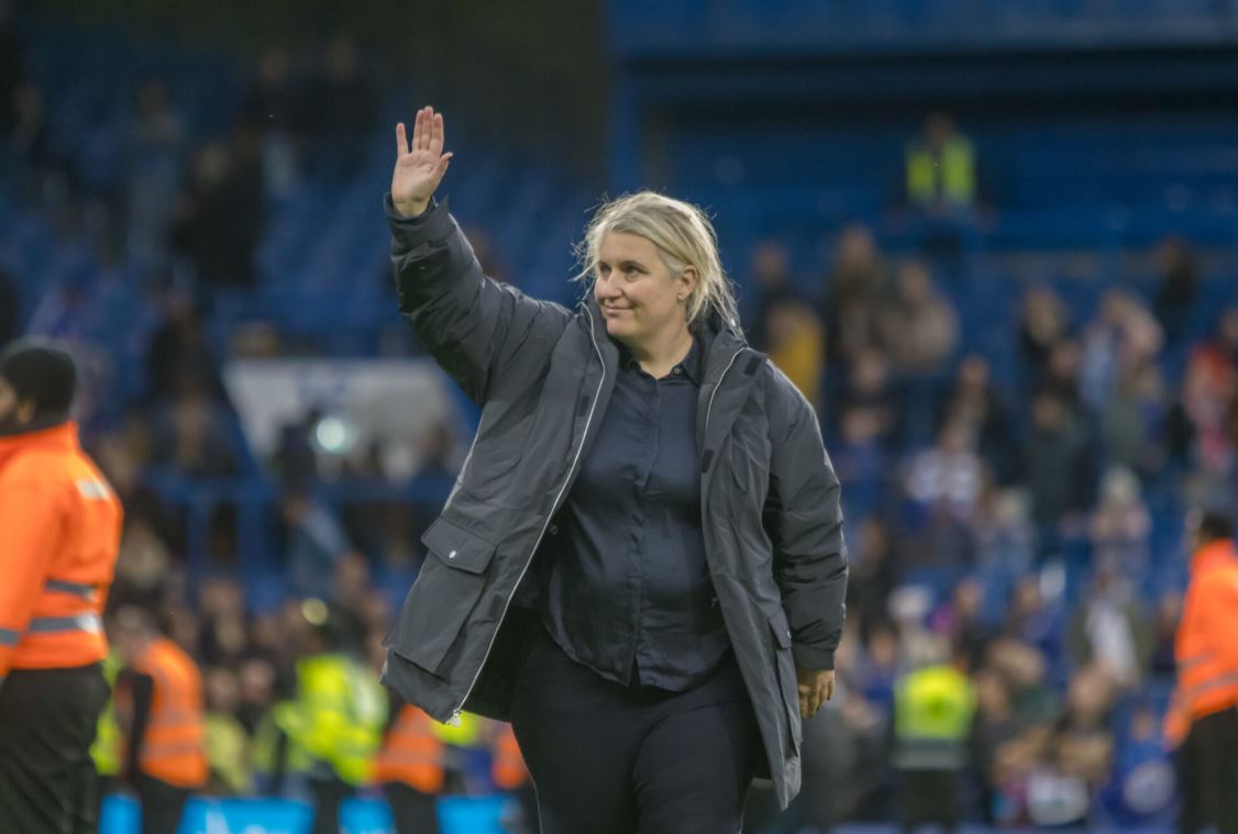 USWNT head coach Emma Hayes is pictured wearing a large overcoat and waving her right hand to a stadium crowd.