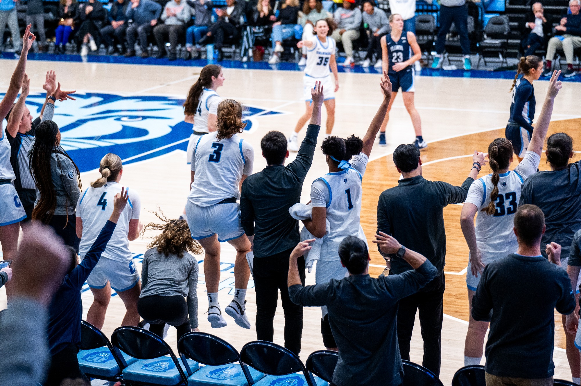 Standing at the top of the key, Columbia guard Abbey Hsu yells and points toward teammate Riley Weiss, who is standing outside the 3-point line. The Columbia bench is visible in the foreground, jumping up and down and making the 3-point hand sign.