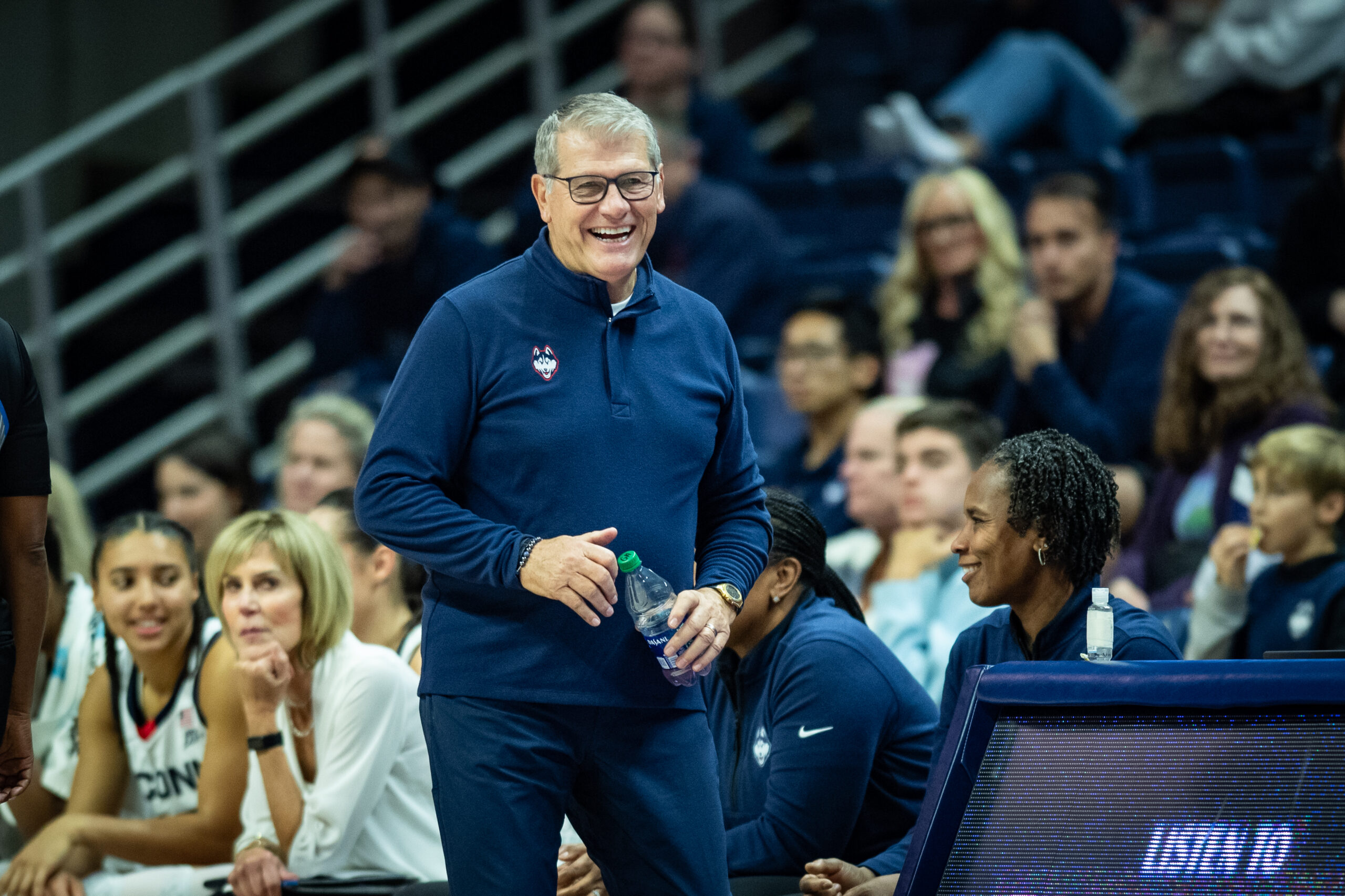 UConn head coach Geno Auriemma smiles on the sidelines. He has a water bottle in his left hand and looks like he's about to use his right hand to unscrew the cap.