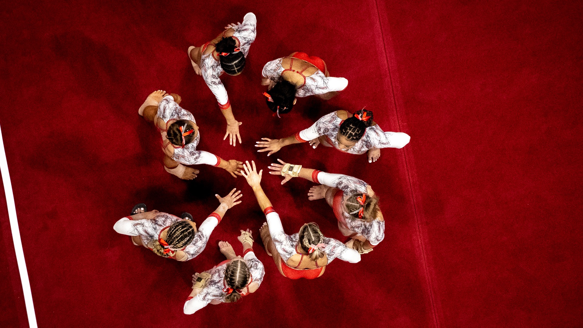 Bird's eye photo of members of Ball State gymnastics gathering in a circle, each with one hand in the middle of the circle.