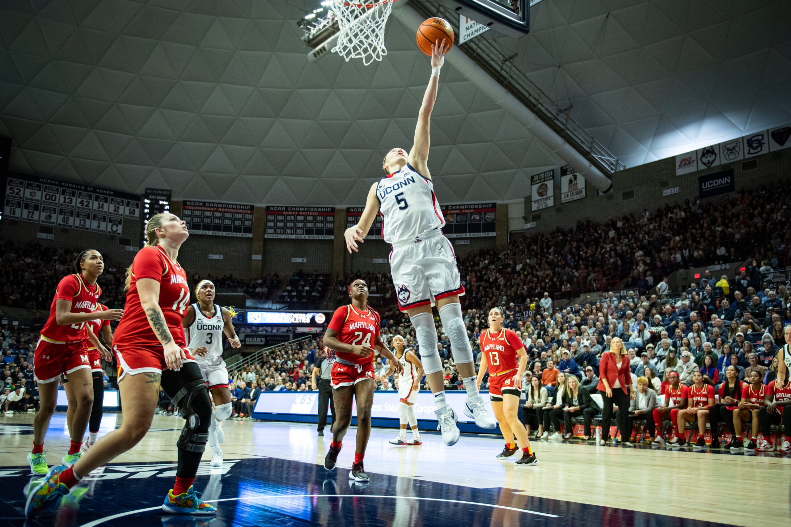 UConn Huskies point guard Paige Bueckers releases a layup midair under the rim, while several Maryland Terrapin defenders look on from behind her