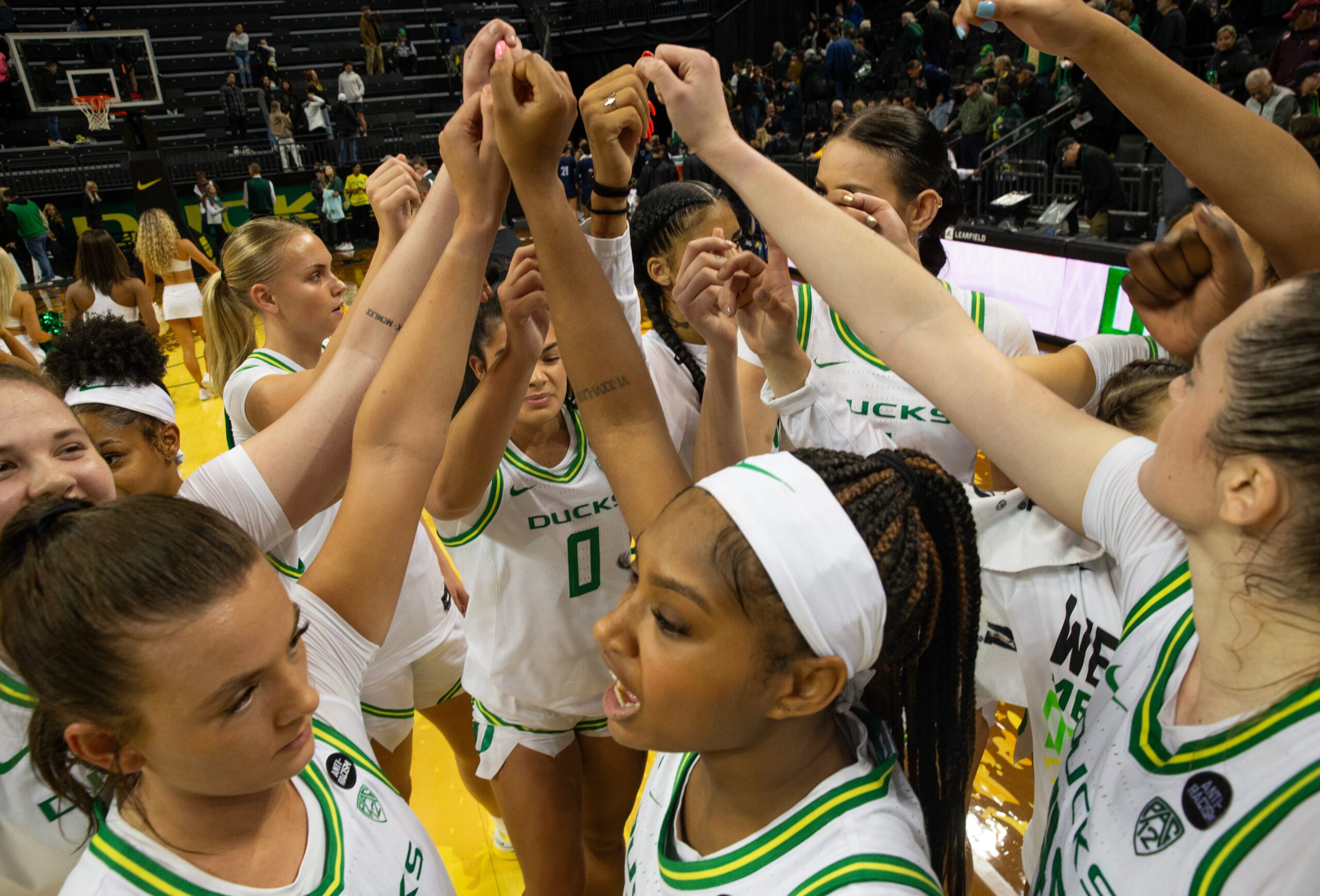 Oregon women’s team come together at mid court in a team huddle. They are wearing their white and green home jerseys.