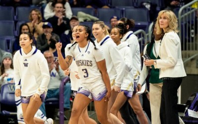 Jayda Noble screams triumphantly and flexes in front of several of her teammates on the bench, celebrating. Tina Langley watches the action from further down the bench on the left.