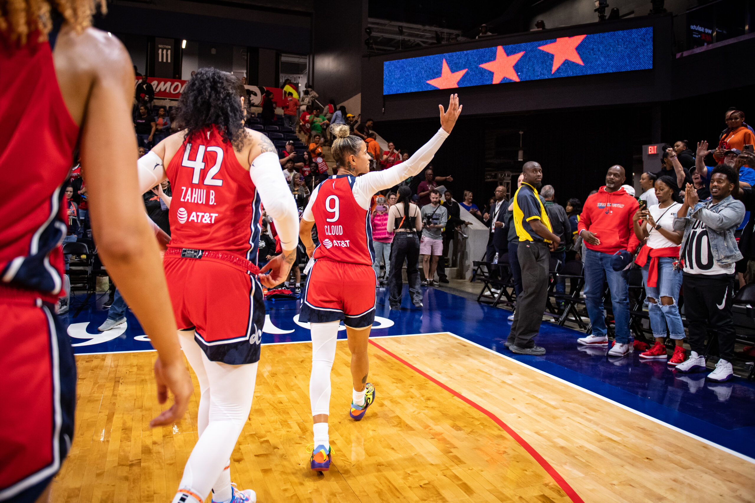 Washington Mystics guard Natasha Cloud walks off the court and looks to her right at fans cheering in the stands. She raises her right arm in acknowledgment.